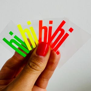 Könnte beinhalten: Eine Hand hält vier transparente Aufkleber mit dem Wort "hi" in Grün, Gelb und Rot. Die Aufkleber haben eine glänzende Oberfläche und werden vor einem weißen Hintergrund gehalten. Die Fingernägel der Person sind rot lackiert.