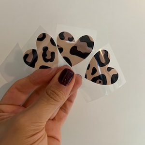 Könnte beinhalten: Drei herzförmige Aufkleber mit Leopardenmuster. Die Herzen sind beige mit schwarzen Flecken und befinden sich auf einer transparenten Folie. Die Aufkleber werden vor einem weißen Hintergrund gehalten.