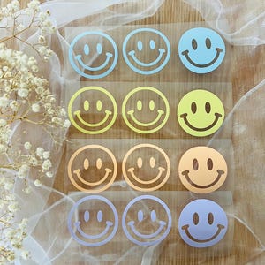 Könnte beinhalten: Neun bunte Smiley-Aufkleber in Blau, Gelb, Orange und Lila. Die Aufkleber sind in drei Reihen zu je drei Stück auf einer transparenten Folie angeordnet. Der Hintergrund umfasst weißen Stoff und eine Holzoberfläche.