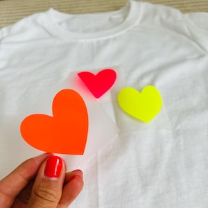 Könnte beinhalten: Weißes T-Shirt mit drei neonfarbenen herzförmigen Bügelbildern in Orange, Rot und Gelb. Die Bügelbilder werden an das Shirt gehalten, was auf ein DIY-Projekt oder eine Individualisierung hindeutet.