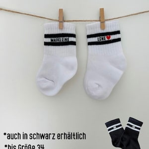 Könnte beinhalten: Ein Paar weiße Socken mit zwei schwarzen Querstreifen und den Namen MADELEINE und LENE mit einem roten Herzsymbol. Ein zweites Paar schwarze Socken mit weißen Streifen und dem Namen LEO ist abgebildet mit dem Text *auch in schwarz erhältlich *bis Größe 34.
