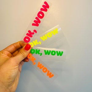 Könnte beinhalten: Eine Hand hält mehrere transparente Aufkleber mit den Worten "OK, WOW" in leuchtenden Farben. Der Text ist in Rot, Gelb, Grün und Orange gehalten. Die Aufkleber sind rechteckig und scheinen aus Vinyl zu sein.
