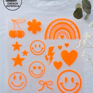 Könnte beinhalten: Ein hellblaues T-Shirt mit einer transparenten Folie mit neonorangen Bügelmotiven. Die Motive umfassen Kirschen, eine Blume, einen Regenbogen, Sterne, Smiley-Gesichter, Herzen, einen Blitz und eine Schleife. Der Text auf der Folie lautet "*auch in anderen Neonfarben erhältlich."
