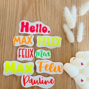 Könnte beinhalten: Eine Auswahl an Namensaufklebern in verschiedenen Neonfarben, darunter Pink, Orange und Grün, auf einem Holzgrund. Die Aufkleber zeigen Namen wie "Max", "Felix" und "Pauline" sowie das Wort "Hello". Dekorative weiße Blumen sind ebenfalls zu sehen.