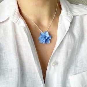 Peut inclure: Un collier délicat en chaîne argentée avec un pendentif en forme de fleur bleu clair. La fleur est en tissu et présente un design superposé et plissé.
