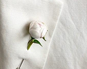 May include: A white peony flower lapel pin with a silver pin backing. The flower has delicate white petals with subtle pink accents. The pin is attached to a white blazer, creating a classic and elegant look.