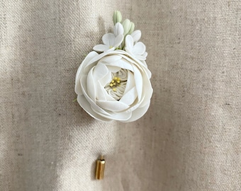 Puede incluir: Un boutonniere de flor de tela blanca con un alfiler dorado. La flor tiene una sola flor blanca con un centro amarillo y flores blancas más pequeñas que la rodean.