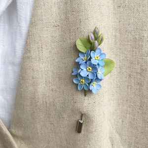 May include: A small bouquet of forget-me-not flowers crafted from polymer clay, featuring a silver pin on the reverse. The flowers are blue with yellow centres and green leaves.