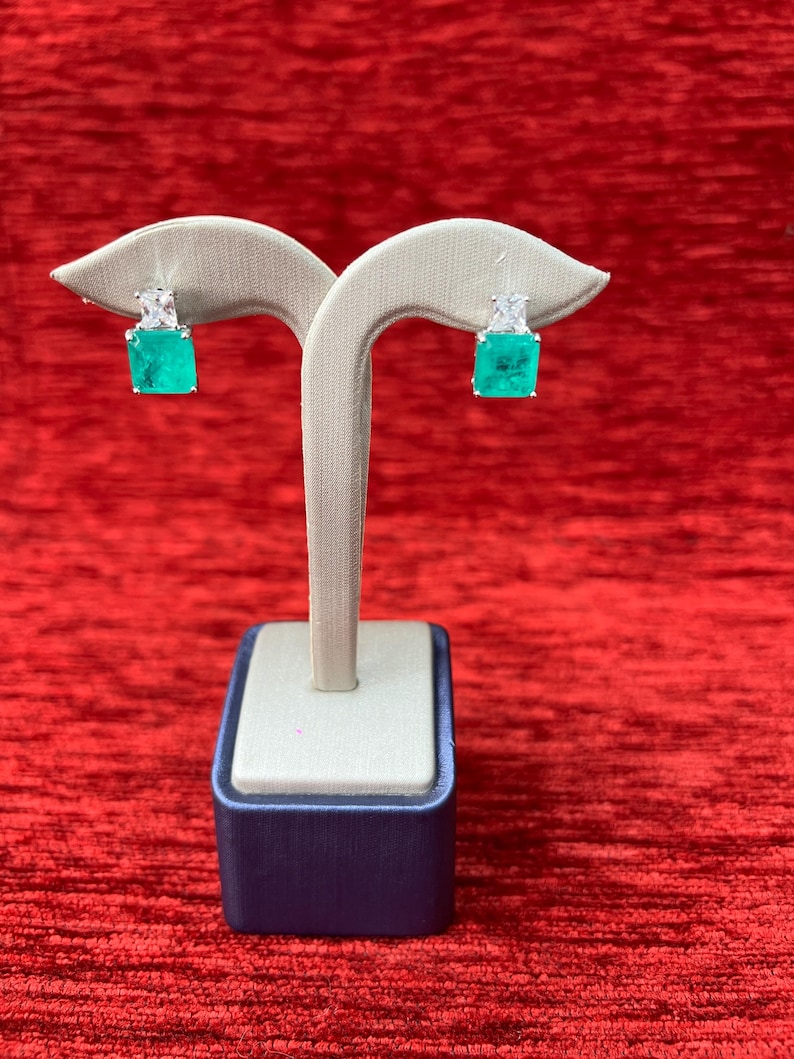Peut inclure: Une paire de boucles d'oreilles carr&eacute;es vert &eacute;meraude avec des accents de diamants, expos&eacute;es sur un support blanc avec une base bleue.