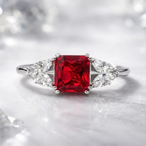 Può includere: Un anello d'argento con una vistosa gemma rossa rettangolare al centro. Ai lati della pietra principale, tre gemme chiare più piccole a forma di pera. L'anello è posto su uno sfondo chiaro e sfocato.