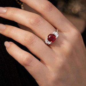 Op de afbeelding: Een zilveren ring met een grote, ovaal geslepen, dieprode edelsteen geflankeerd door twee kleinere, witte edelstenen. De ring zit aan een hand met verzorgde nagels, tegen een vage achtergrond.