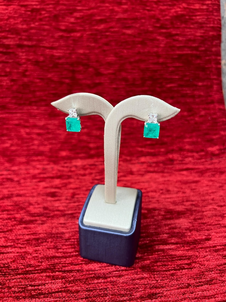 Peut inclure: Une paire de boucles d'oreilles en &eacute;meraude verte et en pierres pr&eacute;cieuses blanches sur un pr&eacute;sentoir. Les boucles d'oreilles sont serties d'argent et ont une forme carr&eacute;e.