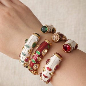 Könnte beinhalten: Eine Sammlung verzierter, farbenfroher Armbänder mit goldenen Akzenten und Schmuckverzierungen. Die Armbänder haben weißen und roten Emaille mit grünen, roten und klaren Edelsteinen. Der Schmuck wird an einem Handgelenk präsentiert.