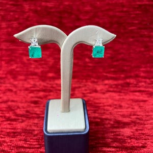 Peut inclure: Une paire de boucles d'oreilles carr&eacute;es vert &eacute;meraude avec des accents de diamants, expos&eacute;es sur un support blanc sur un fond de velours rouge.