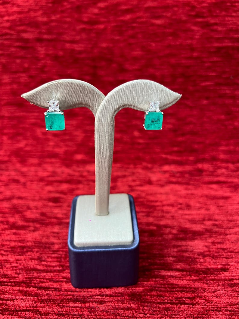Peut inclure: Une paire de boucles d'oreilles carr&eacute;es vert &eacute;meraude avec des accents de diamants, expos&eacute;es sur un support blanc sur un fond de velours rouge.