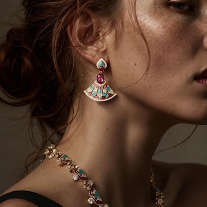 May include: Close-up of a gold-toned earring with a fan-shaped design. The earring features red, turquoise, and white accents, with a pink gemstone and a turquoise heart-shaped top. A matching necklace is also visible.