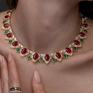 May include: A gold necklace featuring oval-cut red gemstones, emerald-cut green gemstones, and small clear gemstones. The necklace is worn around the neck. A hand with rings is in the foreground.