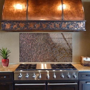 May include: A copper range hood with ornate detailing above a stainless steel gas range. The backsplash has a textured, mottled appearance. A small potted plant in a red pot sits on the countertop.