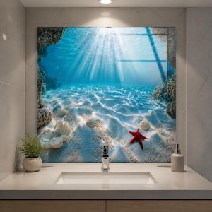 May include: A square wall art piece featuring an underwater ocean scene with a sandy floor, white seashells, and a bright red starfish. Sunlight filters through the clear blue water, creating light rays and ripples on the seabed near rocky formations.
