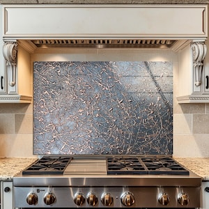 May include: A stainless steel gas range with a decorative backsplash featuring a textured, abstract design in shades of gray and copper. The kitchen also includes white cabinets, a granite countertop, and a wooden knife block.