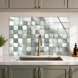 Puede incluir: Un panel rectangular para salpicadero de cocina con una cuadrícula de azulejos cuadrados de nácar iridiscente en tonos blanco, gris y plata. La superficie tiene un acabado brillante y reflectante, situado detrás de un grifo de cocina de acero.
