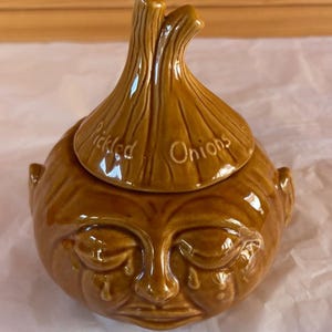May include: A brown glazed ceramic jar shaped like an onion with a matching lid. The lid features the embossed text Pickled Onions. The base is sculpted with a human-like face showing closed eyes and tear-like markings on the cheeks.