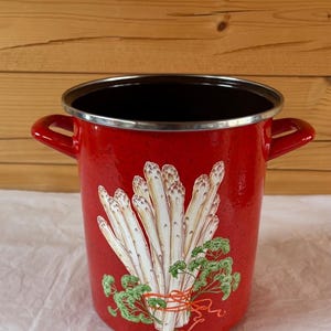 May include: A red enamel cooking pot with a silver rim and two side handles. The pot features a graphic of white asparagus tied with a red ribbon and green parsley. The background is a light wood.