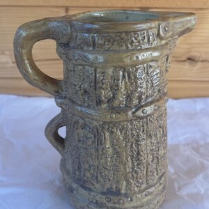 May include: A cylindrical ceramic pitcher with a textured, bark-like surface finish in shades of tan and brown. The vessel features a prominent side handle, a small lower handle, and horizontal bands with circular rivet-like details encircling the body.
