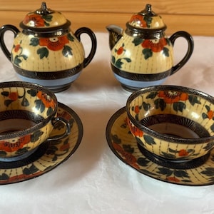 May include: A ceramic tea set featuring a sugar bowl with a lid, a creamer with a lid, and two teacups with matching saucers. Each piece has a cream-coloured base with orange floral patterns, dark green leaves, and black accents with a light blue band.