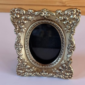 May include: A gold-coloured picture frame with an oval centre opening and a rectangular outer edge. The frame features ornate, raised floral and scrollwork relief patterns throughout its surface, with a dark, recessed inner border.