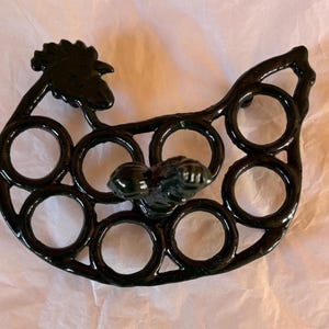May include: A black metal egg holder shaped like a hen. It features eight circular spaces for eggs and a decorative rooster head. This whimsical design is a charming addition to any kitchen.