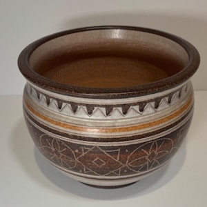 May include: A round ceramic bowl with a brown interior and exterior. The exterior features a decorative pattern of white, orange, and brown bands, along with geometric and floral designs. The rim is a darker brown.