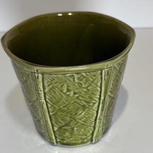 May include: A green ceramic planter with a textured, woven pattern. The planter has a wide, slightly curved rim and a tapered shape. The exterior features vertical bamboo-like accents. The interior is a darker shade of green.