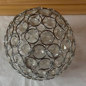 May include: A spherical decorative object with a silver metal frame and clear, faceted glass or crystal accents. The design features interconnected circles, creating a textured, openwork pattern. Likely used for home decor.