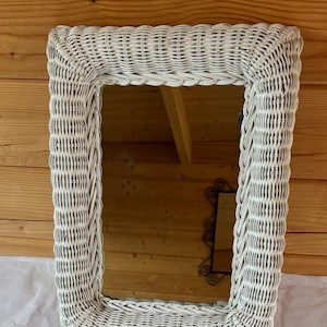 May include: Rectangular white wicker mirror. The frame is made of woven wicker, creating a textured surface. The mirror reflects the interior of a room. The mirror is a decorative item for home decor.
