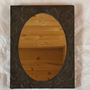 May include: A rectangular mirror featuring a dark, matte-finished frame with an embossed leaf pattern surrounding a central oval reflective surface. The frame has a deep charcoal or black tone, providing a contrast to the clear glass mirror centre.