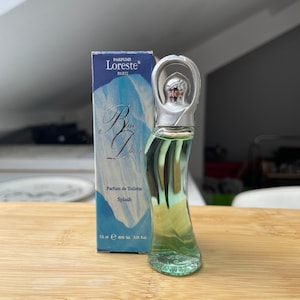 May include: A small, clear glass bottle of blue perfume with a silver cap. The bottle is shaped like a curved, elongated teardrop. The bottle is labelled "Parfums Loreste Paris" and "Blue Dress Parfum de Toilette Splash 7.5 ml e 60% vol 0.25 fl oz".