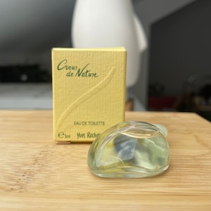 Könnte beinhalten: Eine kleine, transparente Glasparfümflasche und eine passende gelbe Schachtel. Auf der Schachtel steht "Coeur de Nature" und "Eau de Toilette". Die Flasche ist oval geformt und enthält eine hellfarbene Flüssigkeit. Die Schachtel weist 5 ml Parfüm auf.