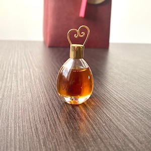 Può includere: Un piccolo flacone di profumo trasparente con tappo dorato e manico a forma di cuore. La bottiglia è riempita con un liquido marrone dorato e reca la scritta "Secret de Venus". La bottiglia è su una superficie di legno.