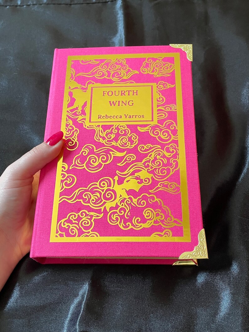 Op de afbeelding: Een roze hardcover boek met gouden accenten en een gouden ontwerp van wolken en een draak. De titel van het boek is "FOURTH WING" door Rebecca Yarros.
