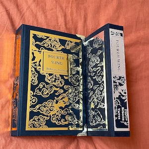 Op de afbeelding: Een hardcover boek met een zwarte omslag en gouden letters. De titel is "Fourth Wing" door Rebecca Yarros. De omslag heeft een gouden ontwerp van wolken.
