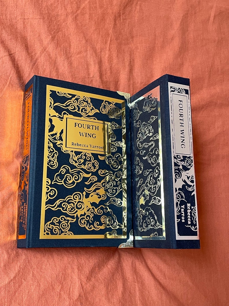 Op de afbeelding: Een hardcover boek met een zwarte omslag en gouden letters. De titel is "Fourth Wing" door Rebecca Yarros. De omslag heeft een gouden ontwerp van wolken.