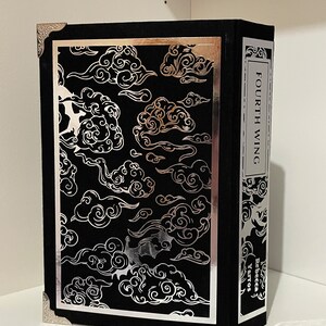 Op de afbeelding: Een zwart boek met een zilveren rand en een zilveren ontwerp van wolken op de voorkant. De titel "FOURTH WING" is in zilver op de rug gedrukt. De naam van de auteur, "Rebecca Yarros", is in zilver op de achterkant gedrukt.