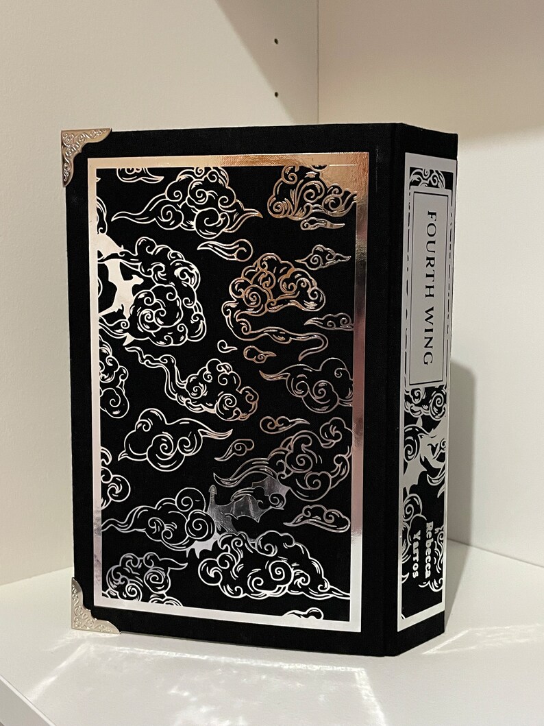 Op de afbeelding: Een zwart boek met een zilveren rand en een zilveren ontwerp van wolken op de voorkant. De titel "FOURTH WING" is in zilver op de rug gedrukt. De naam van de auteur, "Rebecca Yarros", is in zilver op de achterkant gedrukt.