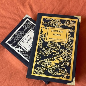 Può includere: Due libri rilegati in tela nera con dettagli dorati e un motivo di nuvole dorate sulla copertina. Il libro in cima ha il titolo "FOURTH WING" e l'autore "Rebecca Yarros" stampato in oro sulla copertina.
