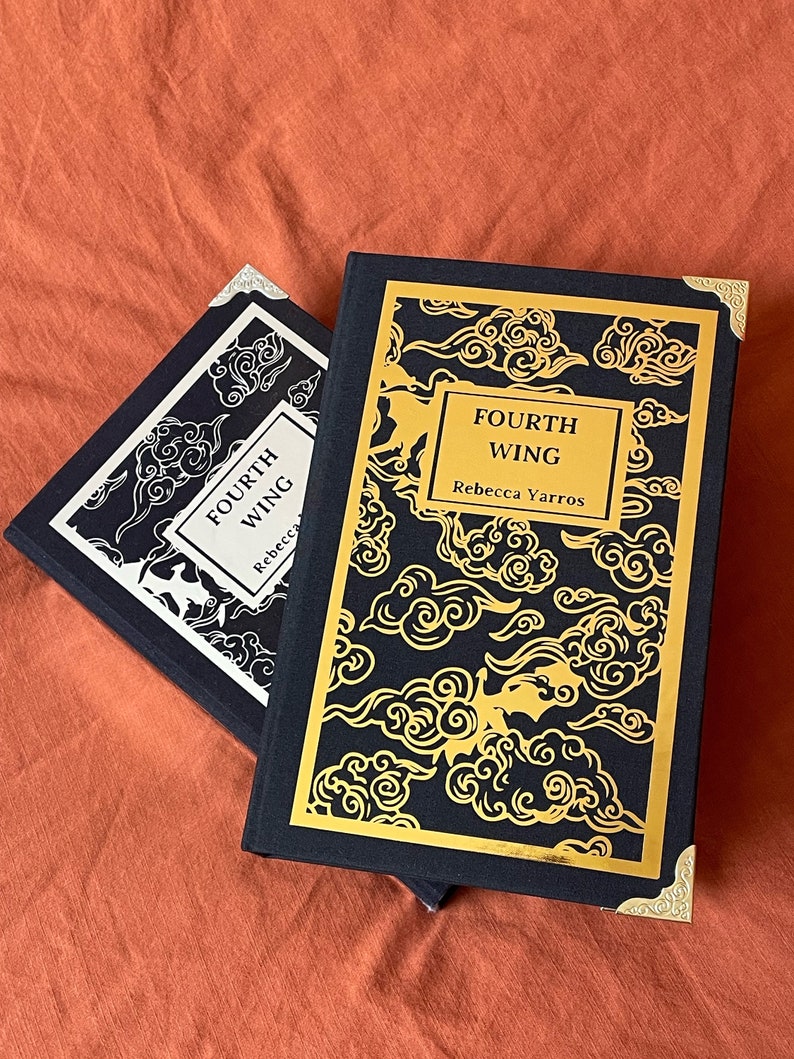 Op de afbeelding: Twee zwarte hardcover boeken met gouden accenten en een gouden wolkpatroon op de omslag. Het bovenste boek heeft de titel "FOURTH WING" en de auteur "Rebecca Yarros" staat in goud op de omslag gedrukt.
