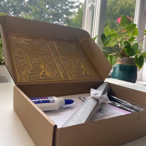 Pu&ograve; includere: Una scatola di cartone marrone con un nastro bianco e un rotolo di carta all'interno. La scatola &egrave; aperta e il contenuto &egrave; visibile. La scatola &egrave; appoggiata su una superficie bianca. La scatola ha un design decorativo all'interno del coperchio.