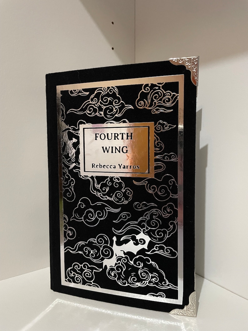 Op de afbeelding: Een zwart, met fluweel gebonden hardcover boek met zilveren foliedetails. De titel "FOURTH WING" en de auteur "Rebecca Yarros" zijn in zilveren folie op de voorkant gedrukt.