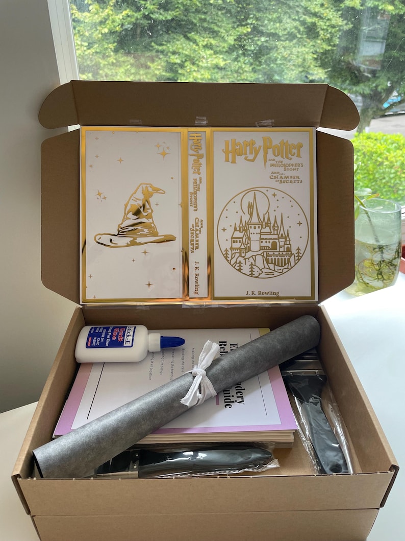 Pu&ograve; includere: Una scatola di cartone aperta piena di forniture per un progetto di ricamo a tema Harry Potter. La scatola contiene due copertine di libri in foglia d'oro, un rotolo di tessuto, una guida, una bottiglia di colla e due aghi da ricamo neri.