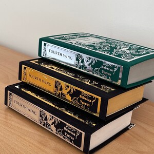 Op de afbeelding: Drie hardcover boeken gestapeld op elkaar. Het bovenste boek is groen met zilveren letters en de woorden "Fourth Wing" op de rug. Het middelste boek is zwart met gouden letters en de woorden "Fourth Wing" op de rug. Het onderste boek is zwart met zilveren letters en de woorden "Fourth Wing" op de rug. Alle drie boeken hebben de naam van de auteur, "Rebecca Yarros", op de rug gedrukt.